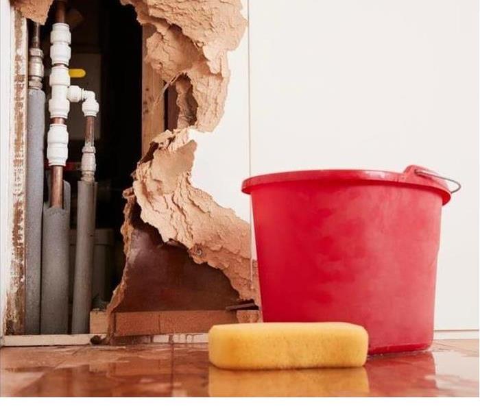 Picture of a wall opened up to see plumbing, with bucket and sponge on wet floor